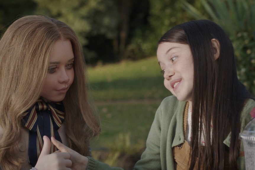 A blonde human-size AI doll sits at a park bench with a young white girl with long dark hair.