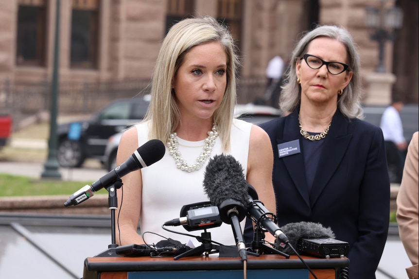 A woman wearing a white singlet and pearl necklace speaks into microphones at a lectern