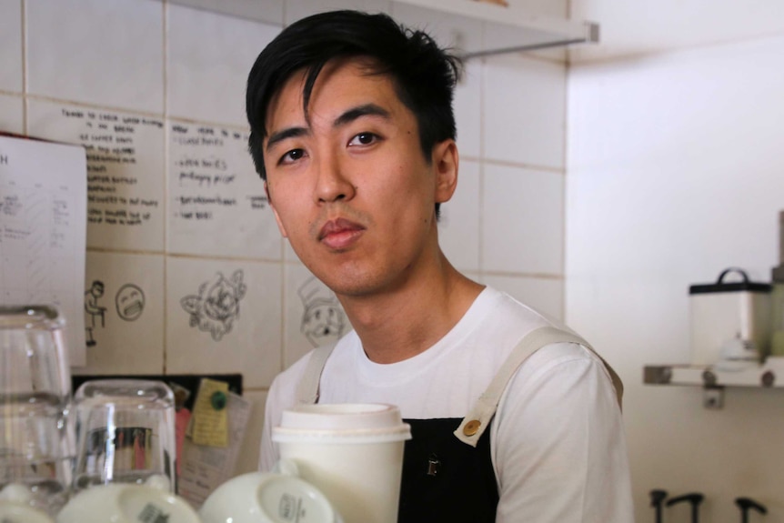 A cafe owner wearing an apron stands behind a barista coffee machine in a cafe.
