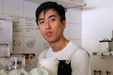 A cafe owner wearing an apron stands behind a barista coffee machine in a cafe.