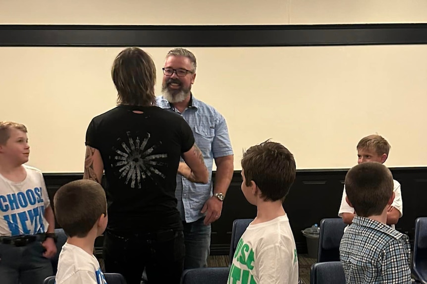  Keith Urban has back to camera, shaking hands with a smiling man, grey beard, glasses, kids look on.