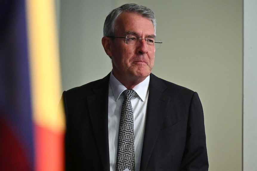 a man wearing glassed stands next to a flag looking sideways