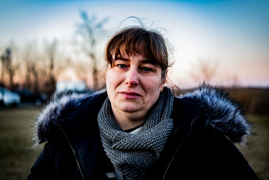 A woman in a fur trimmed coat looking frightened while standing in a field at sunset