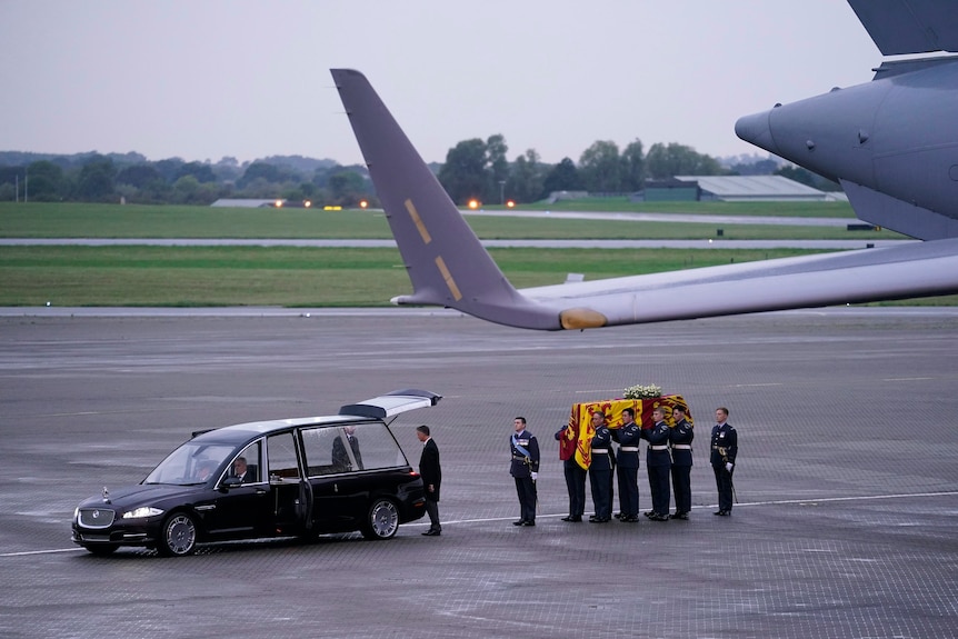 The flag-draped coffin of Queen Elizabeth II is carried across a tarmac to a waiting hearse, with a plane wing in the foreground