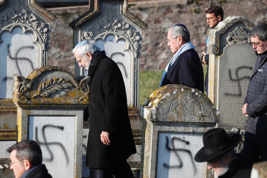 Men in black coats walk with their heads down among tombstones with swastikas painted on them.