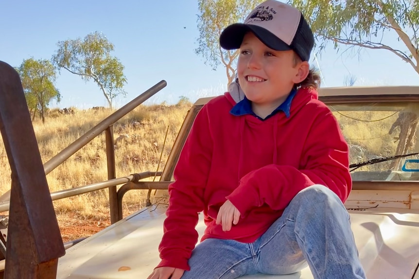 boy wearing red long-sleeve shirt and cap, smiling in front of rural cattle station backdrop