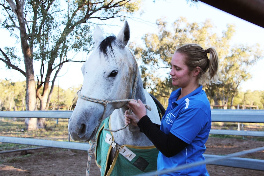 A woman in a blue work shirt adjusts the bridle on a horse.