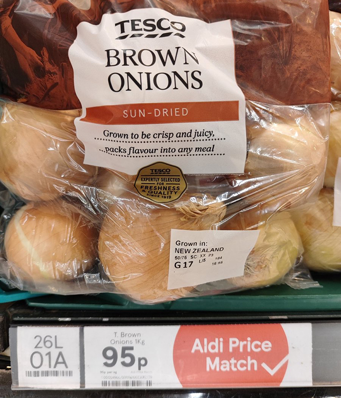 A bag of brown onions with Tesco branding and a shelf price point label that notes an Aldi price match