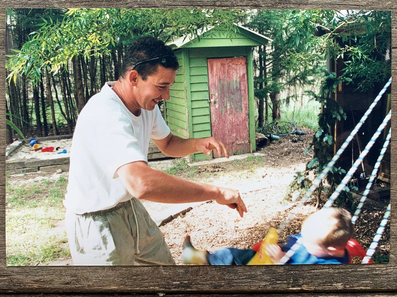 A man wearing Tshirt and shorts smiles as he pushes a child on a swing, in a yard in front of a red shed