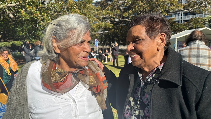 Two older Aboriginal women smile at each other at an outside event in a green park.