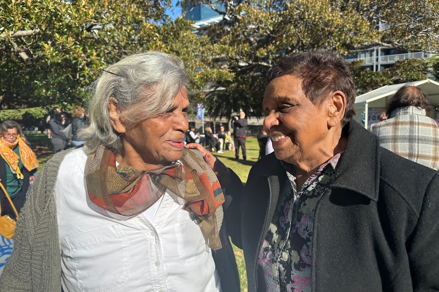 Two older Aboriginal women smile at each other at an outside event in a green park.
