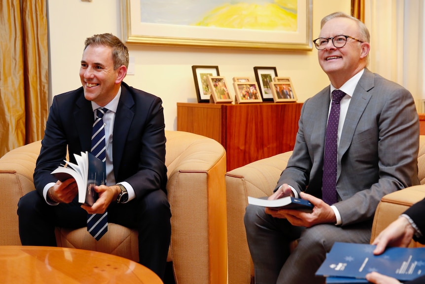 Jim Chalmers and Anthony Albanese sit on a couch holding the budget booklets. They are smiling.