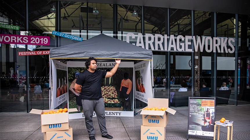 A man with dark curly hair and t-shirt stands with marquee and artwork out front of Carriageworks arts venue.
