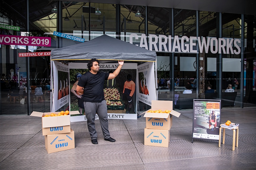 A man with dark curly hair and t-shirt stands with marquee and artwork out front of Carriageworks arts venue.