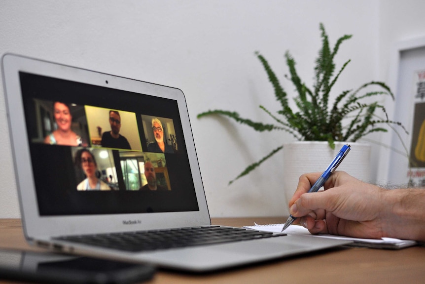 A laptop on a table. On its screen is a video conference call with five people. A hand sits on the table holding a pen.