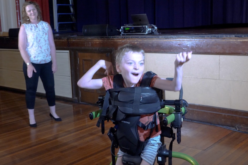 A young boy in a wheelchair waving his arms above his head as his mum watches on.