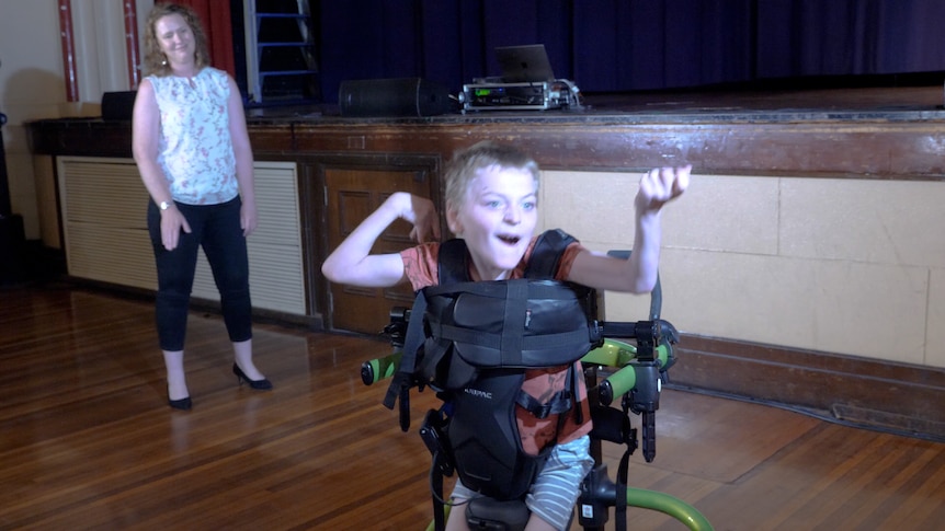 A young boy in a wheelchair waving his arms above his head as his mum watches on.