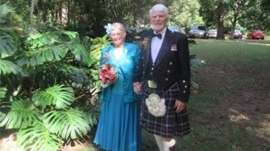 elderly woman in blue dress and elderly man in Scottish kilt hold hands surrounded by rainforest