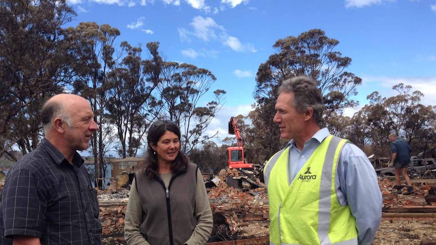 Tim and Tammy Holmes stand in front of house ruins with the head of Aurora Energy Peter Davis.