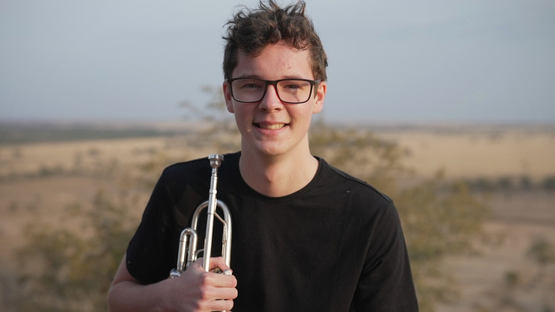 Malcolm smiles to camera holding a trumpet in the outback somewhere near Longreach.