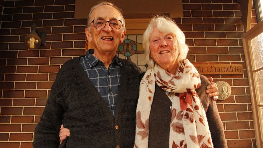 An older man stands with his arm around his wife, in front of a brick house.