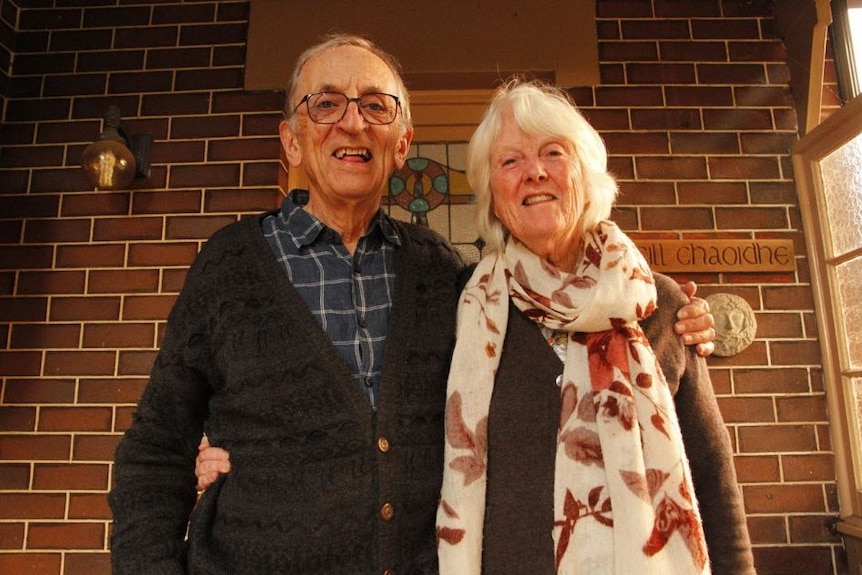 An older man stands with his arm around his wife, in front of a brick house.