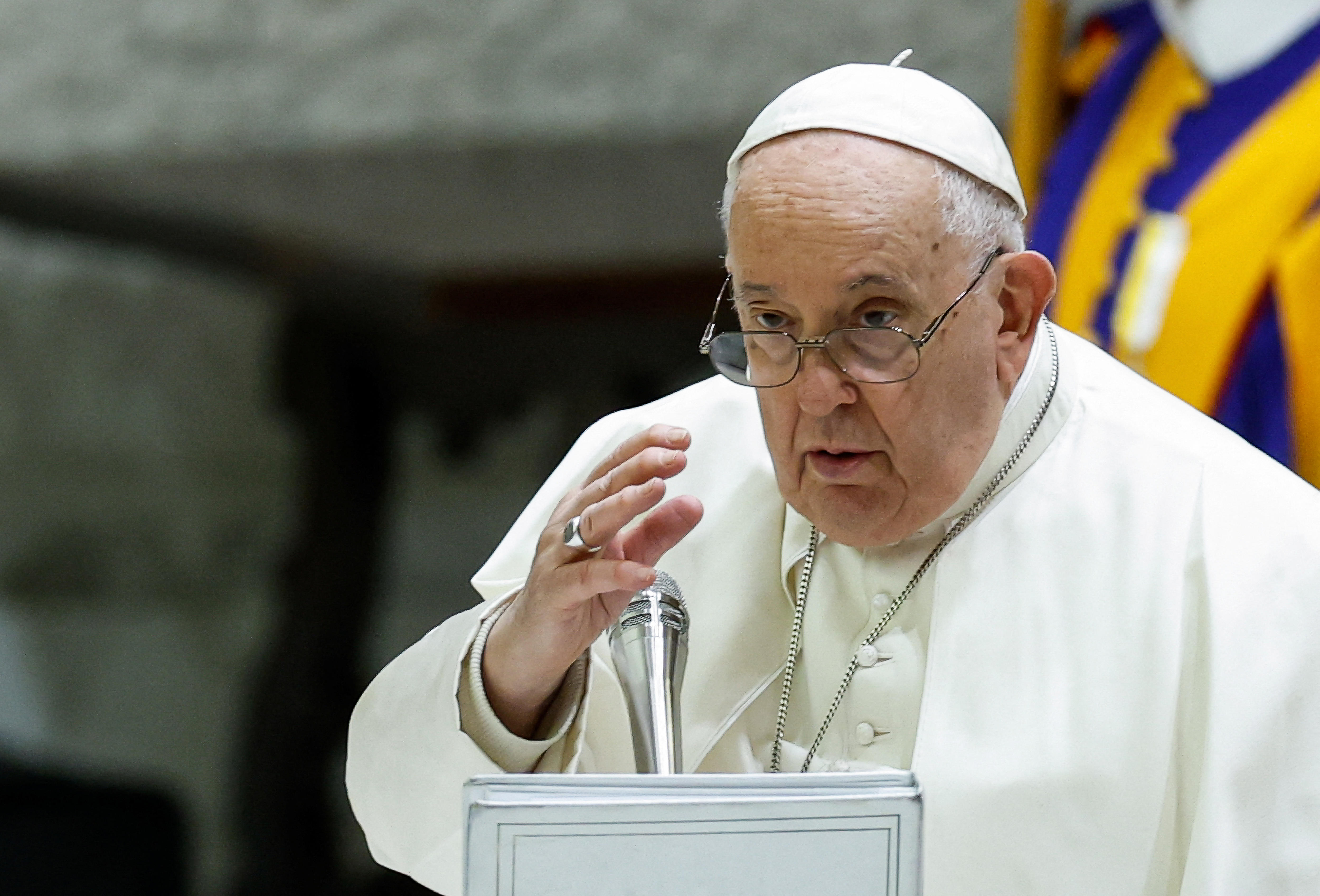 An elderly man in white clothing and a skull cap gestures as he speaks into a silver microphone.