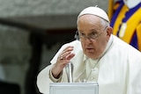 An elderly man in white clothing and a skull cap gestures as he speaks into a silver microphone.