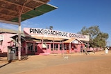 A single-storey building stands under a blue sky and red dirt with a sign saying Pink Roadhouse Ooodnadatta.