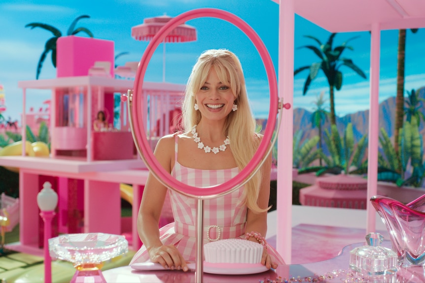 Margot Robbie smiles while holding an oversized plastic brush, wearing a pink and white gingham dress