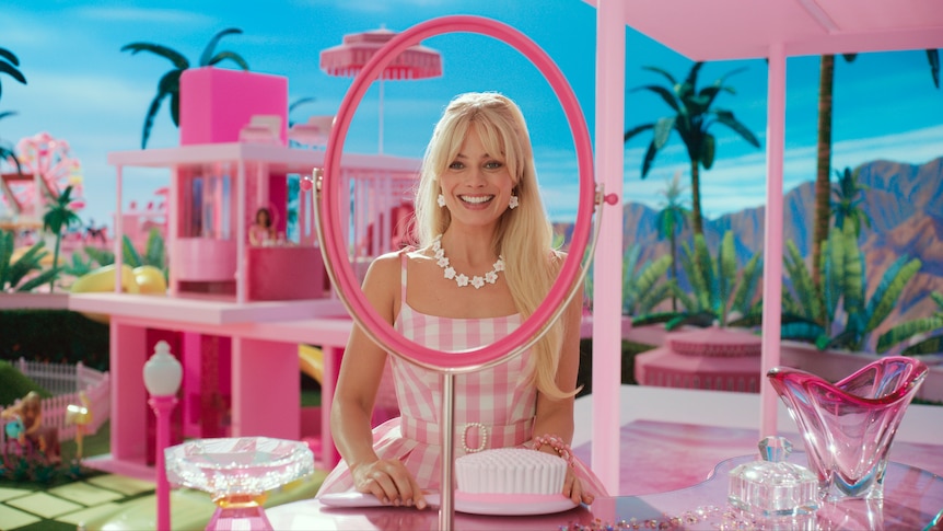 Margot Robbie smiles while holding an oversized plastic brush, wearing a pink and white gingham dress