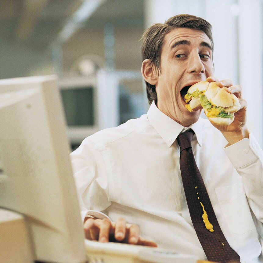 Man sitting at computer eating a roll while working and mustard is dribbling down his tie
