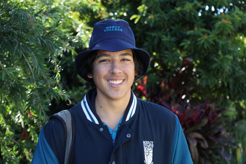 Un adolescente varón sonriente con uniforme escolar, un sombrero azul con Mercy College sonríe a la cámara.