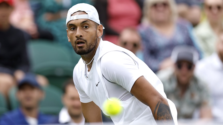 Nick Kyrgios, wearing a backwards cap, watches a tennis ball go over the net at Wimbledon.