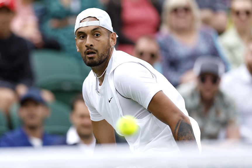 Nick Kyrgios, wearing a backwards cap, watches a tennis ball go over the net at Wimbledon.