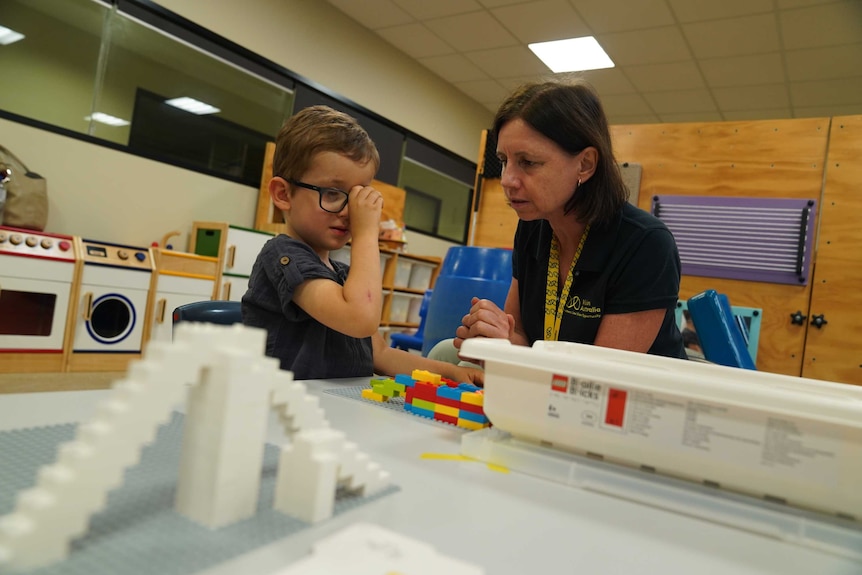 A young boy holds a Lego block close to his face, while a woman leans down beside him. They're in a classroom-type setting.
