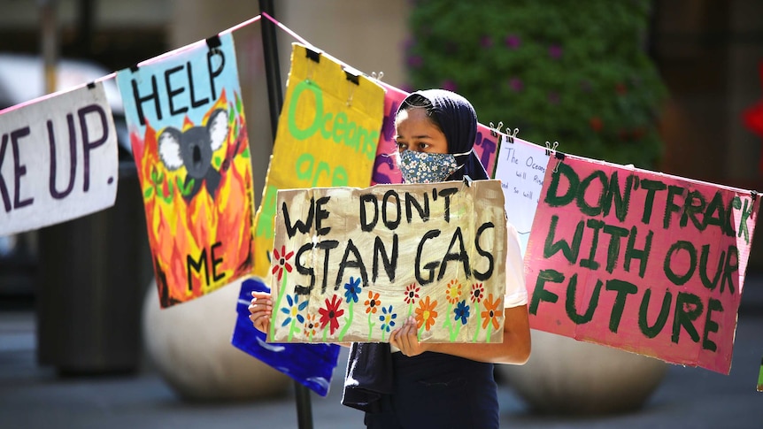 A young girl stands in front of the washing line of signs holding her own, which reads "we don't stan gas"