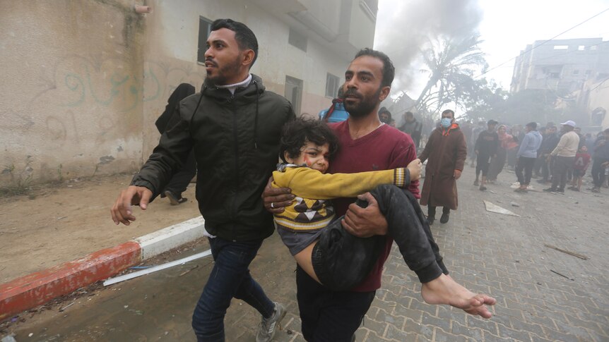Man carries young children wearing yellow jumper with blood on their face. 