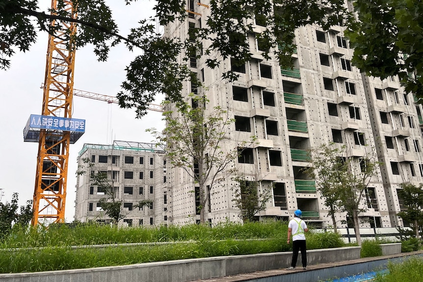 A man looks at a yellow crane next to a grey apartment building.