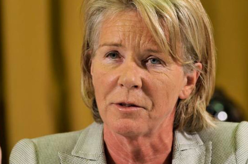 A woman with short blonde hair in a green suit uses her right hand to gesture while talking.