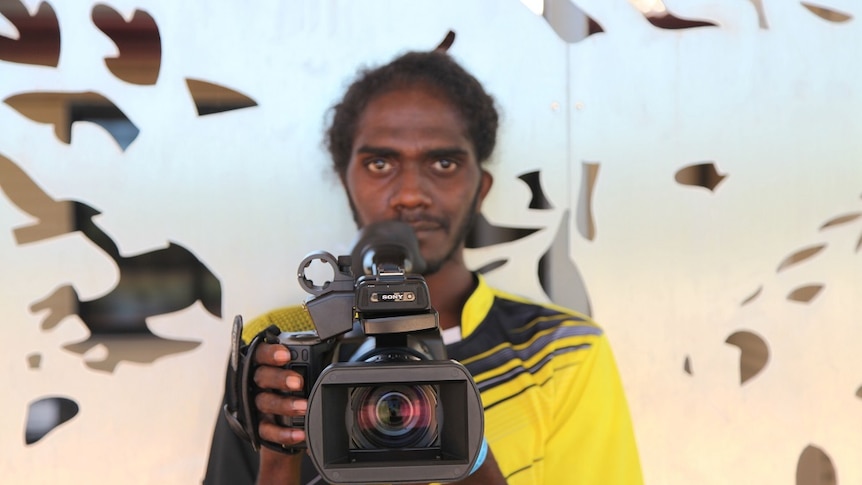 A young Aboriginal man in a yellow shirt stands with a camera held out in front of him