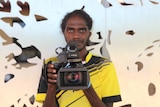 A young Aboriginal man in a yellow shirt stands with a camera held out in front of him