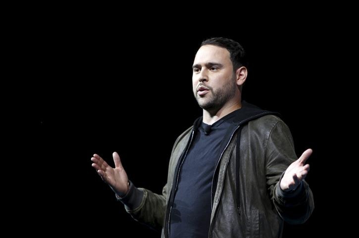 Scooter Braun speaks holding his hands up as he stands against a black background. He wears a leather jacket and black t-shirt.