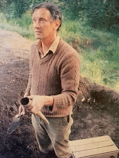 Alex Podolinsky stands in a trench holding a trowel and hollow cow horn.