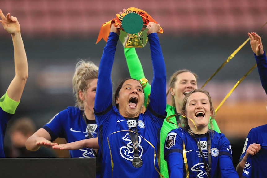 Sam Kerr lifts a trophy above her head with her mouth wide open, wearing a blue kit