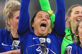Sam Kerr lifts a trophy above her head with her mouth wide open, wearing a blue kit