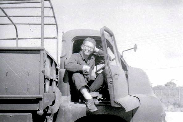Black and white photo shows a young man sitting in the drivers seat of a truck smiling.