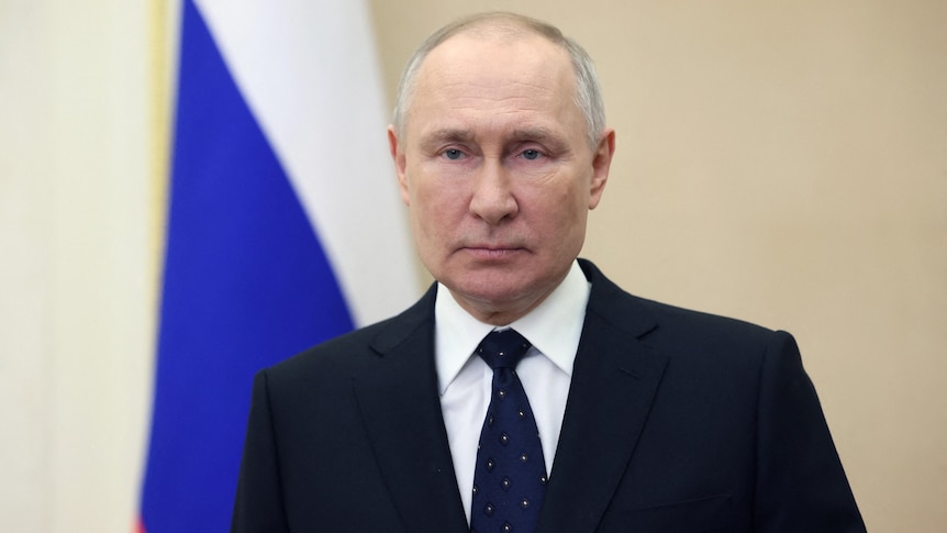 Close up of suited Vladimir Putin with Russian flag in background.