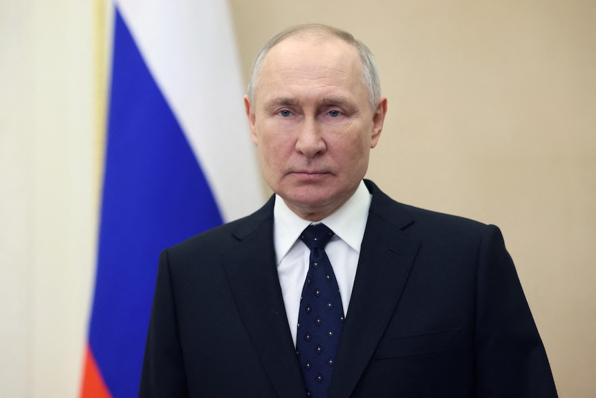 Close up of suited Vladimir Putin with Russian flag in background.