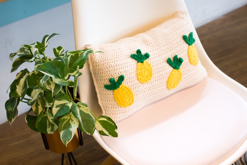 A photo of a yellow and green plant next to a cream chair with cushion crocheted with pineapples